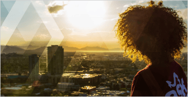 A person is sitting on a mountain and looking at the sun and there are buildings close to the mountains